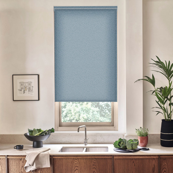 Blue roller blind covers most of a window, blocking light; kitchen sink with chrome faucet on marble countertop, wooden cabinets below, nearby potted plants, bowl of greens and framed sketch.