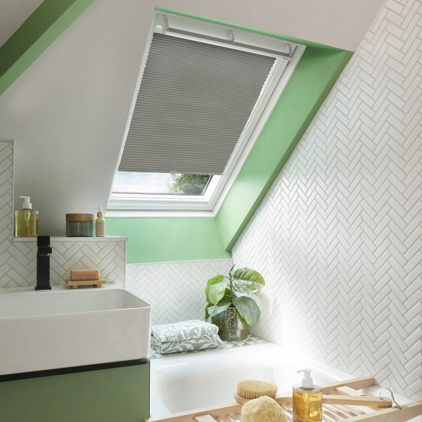 Grey pleated skylight blind, partially lowered and filtering daylight, in a green-accented attic bathroom with white herringbone tiles, sink and bathtub, folded towel and potted plant on ledge.