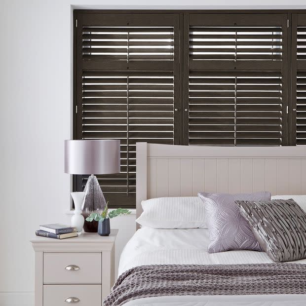 Dark wooden horizontal slatted shutters spanning the window behind the bed, slats partially tilted to filter thin light; bedroom setting with pale headboard, nightstand, lamp, pillows and soft lavender accents.