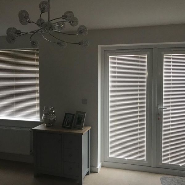 Horizontal venetian blinds on a window and patio door, closed and filtering dim daylight; living-room corner with a small cabinet, owl ornament, two photos, and chandelier.