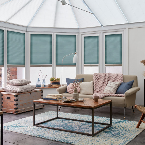 Teal cellular blinds covering tall conservatory windows, fully lowered pleated honeycomb panels filtering soft daylight; setting a bright conservatory living room with beige sofa, wooden coffee table, and textured rug.