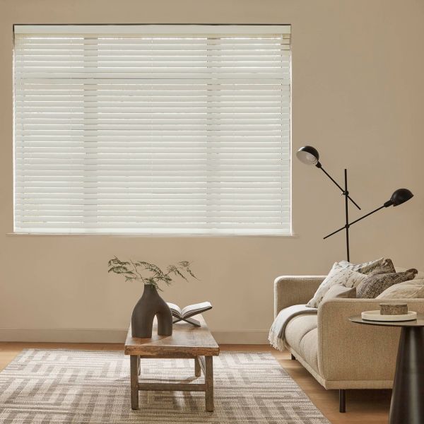 White horizontal blinds fully lowered and closed, softly filtering daylight through slats; beige living room with a neutral sofa, wooden coffee table, patterned rug and black floor lamp.