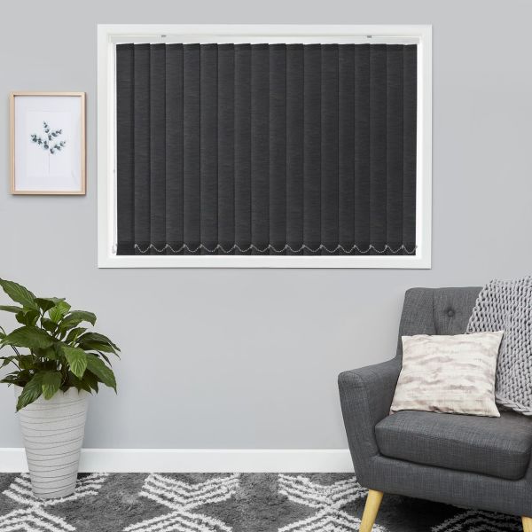Vertical dark grey blinds cover a white-framed window, fully closed with beaded chain weights; pale grey living-room wall flanked by a potted plant left and an upholstered armchair right.