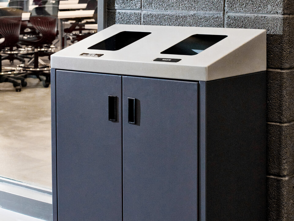 double waste and recycling collection container with a sloped metal lid and grey metal body with hinged front doors in a high school hallway