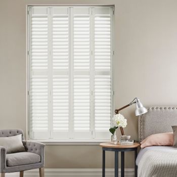 White plantation shutters — four hinged louvered panels covering the window — closed, slats filtering bright daylight. Neutral-toned bedroom with upholstered headboard, bedside table and lamp, armchair nearby.