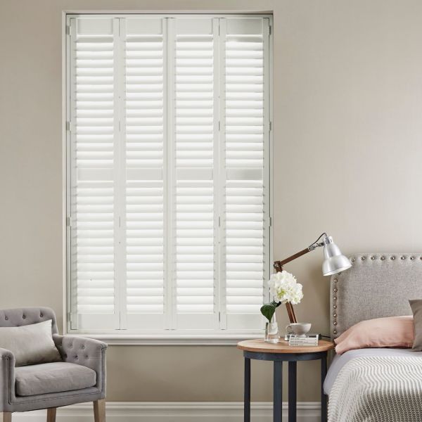 White plantation shutters — four hinged louvered panels covering the window — closed, slats filtering bright daylight. Neutral-toned bedroom with upholstered headboard, bedside table and lamp, armchair nearby.