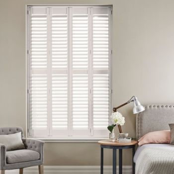White full-height plantation shutters with four framed panels and horizontal louvers, closed and filtering bright daylight; set in a neutral-toned bedroom corner with upholstered bed, side table, lamp, and armchair.