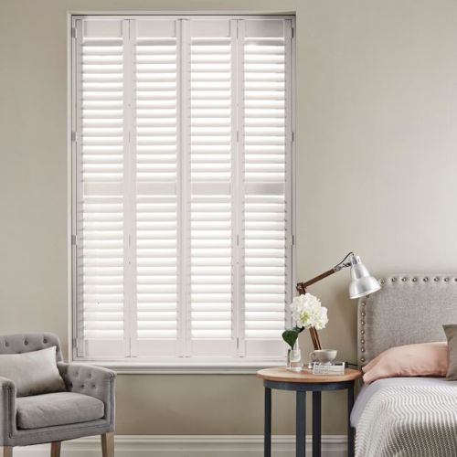White full-height plantation shutters with four framed panels and horizontal louvers, closed and filtering bright daylight; set in a neutral-toned bedroom corner with upholstered bed, side table, lamp, and armchair.