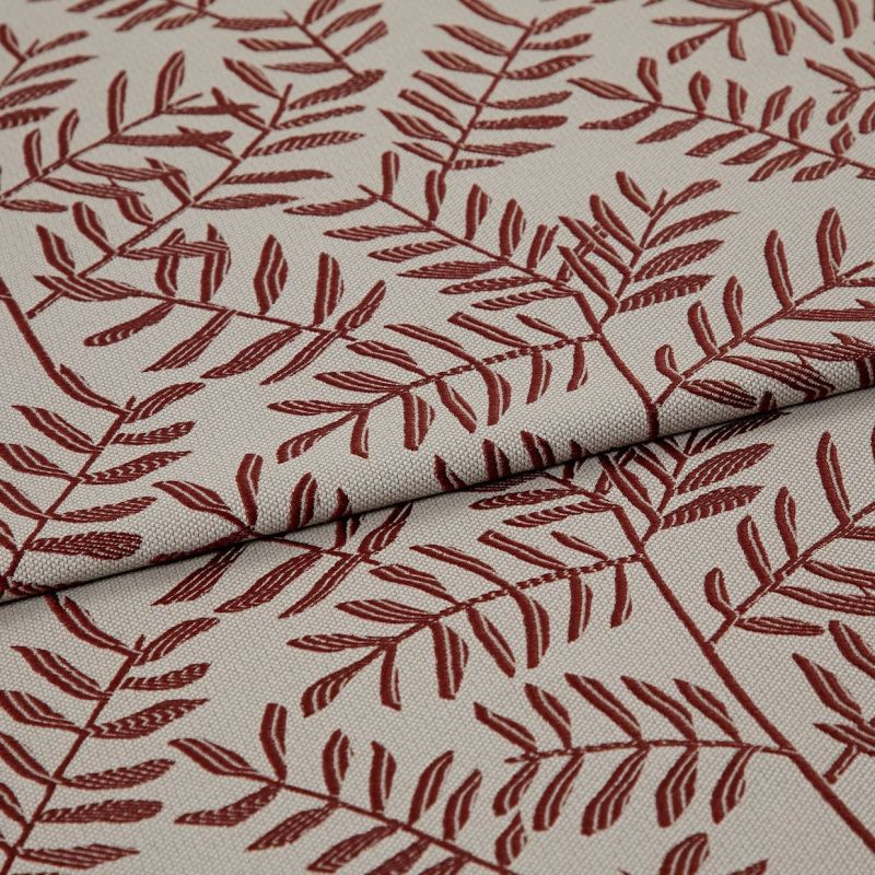 Woven beige upholstery fabric patterned with rust-red fern branches, folded and overlapped across the frame, close-up studio-lit view highlighting the woven texture and printed details.