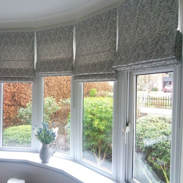 Leaf-patterned fabric Roman blinds across a bay of windows, partially lowered with visible pull chains, softly filtering daylight; sill holds a jug of flowers, view of manicured shrubs outside.