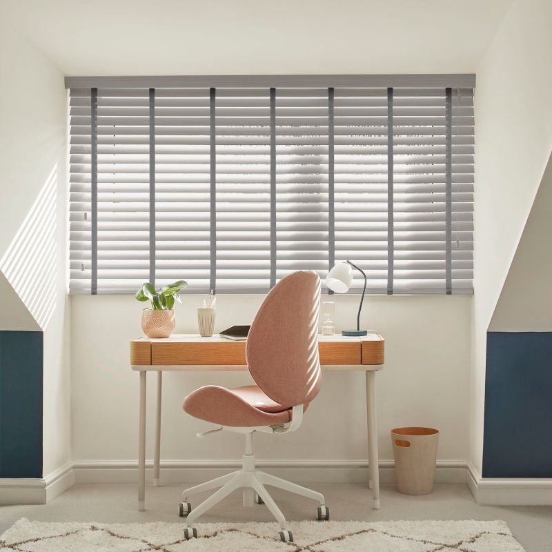 Grey horizontal Venetian blinds, full-width with vertical tapes and tilted slats, filtering sunlight and casting striped shadows over an attic study nook with wooden desk, pink swivel chair, lamp, plant.