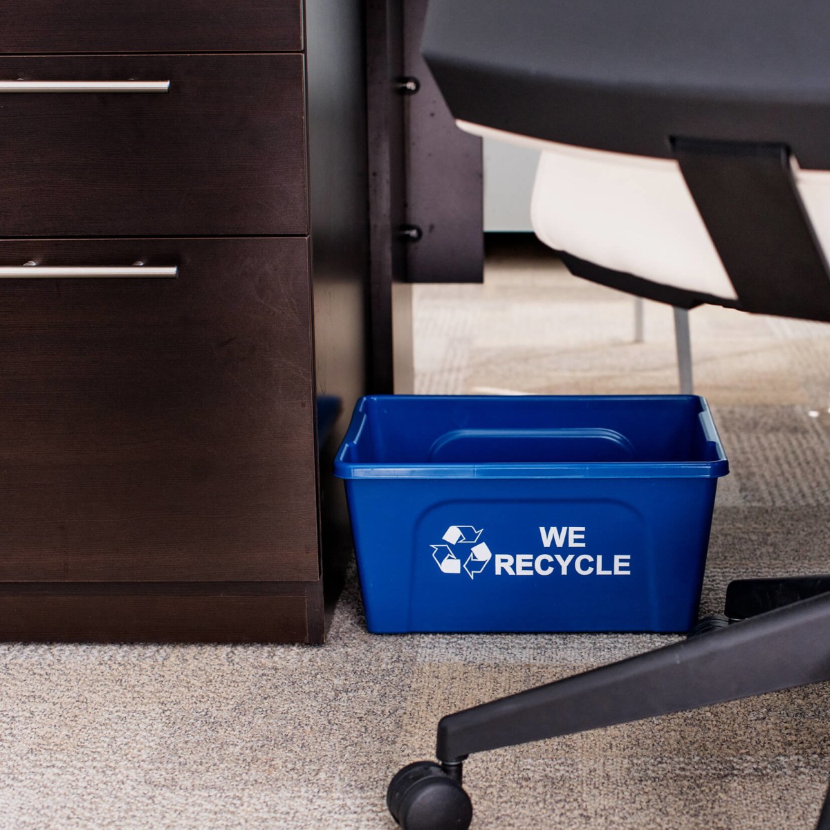 small blue plastic recycling bin with we recycle printed on the side sitting on the floor under an office desk