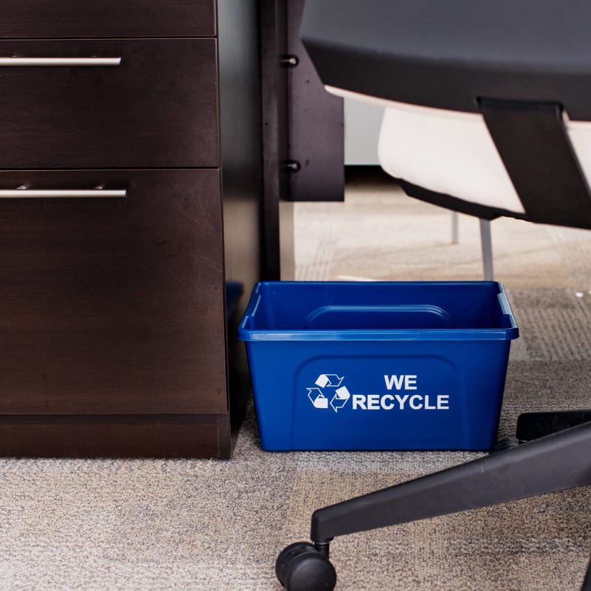 small blue plastic recycling bin with we recycle printed on the side sitting on the floor under an office desk