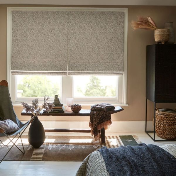 Textured beige Roman shades, lowered to mid-window with a slight bottom gap, softly filtering daylight; set above a bench in a neutral-toned bedroom nook with woven baskets.