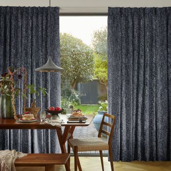 Dark gray textured pleated floor-length curtains, drawn aside at center to frame the opening; open, admitting bright daylight and a lush backyard view beyond a dining table.