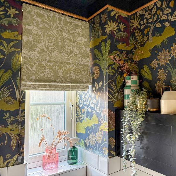 A patterned fabric Roman blind, lowered over a frosted window, filters daylight; botanical navy-and-gold wallpaper frames a tiled bathroom sill topped with glass vases and cascading faux foliage.