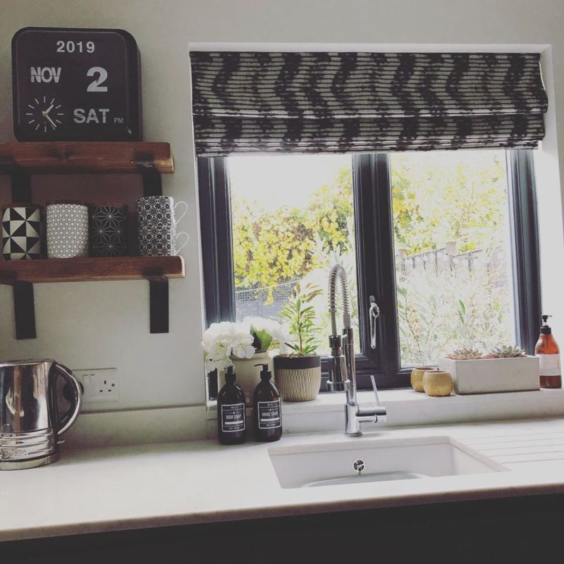 Patterned fabric Roman blind, partially lowered across a black-framed double window, filtering bright daylight; stainless spring-tap over kitchen sink, potted plants, soap bottles and wooden mug shelves nearby.
2019
NOV
2
SAT
PM
DISH SOAP
HAND SOAP