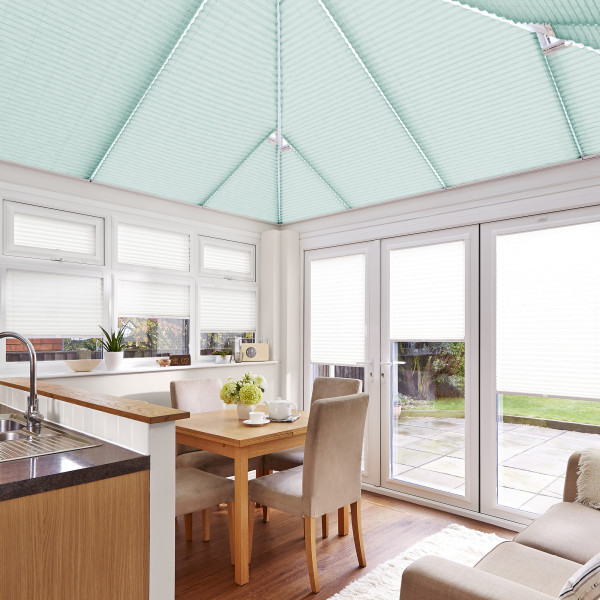 Seafoam pleated roof blinds cover the pitched conservatory ceiling; white pleated blinds cover windows and doors, filtering soft daylight into a bright kitchen-dining conservatory with wooden table and garden view.