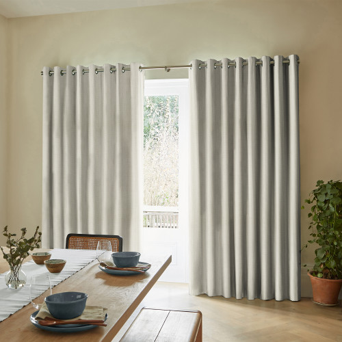 Grommet-top light-gray heavy curtains, floor-length and slightly parted at the center, filtering bright daylight from a glass door; dining room with wooden table settings, bench seating, and a potted plant.