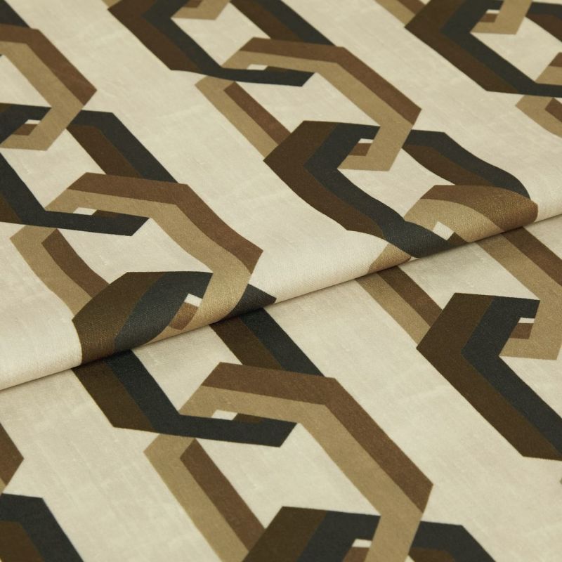 Cream fabric sample printed with repeating brown and black interlocking geometric shapes; folded across the center; close-up studio shot with soft, even lighting on a neutral background.