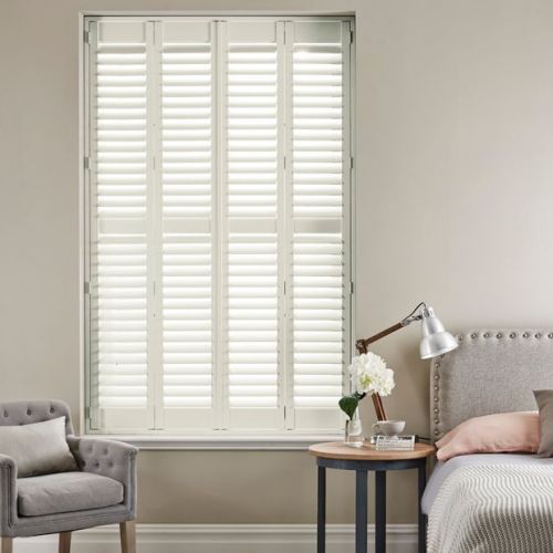 White plantation shutters covering a tall window, closed with angled louvers filtering soft daylight; bedroom setting with upholstered bed, round side table with lamp and flowers, armchair, and neutral walls.