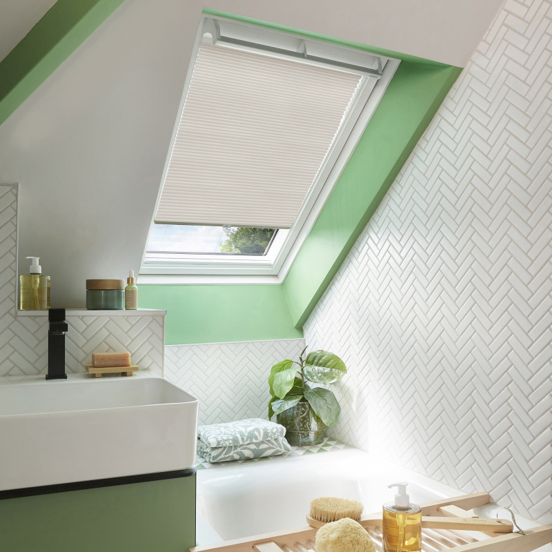 Cream pleated cellular blind lowered over a skylight, filtering daylight; set in a bright attic bathroom with mint-green window frame, white herringbone tiles, bathtub, sink, plant and toiletries.