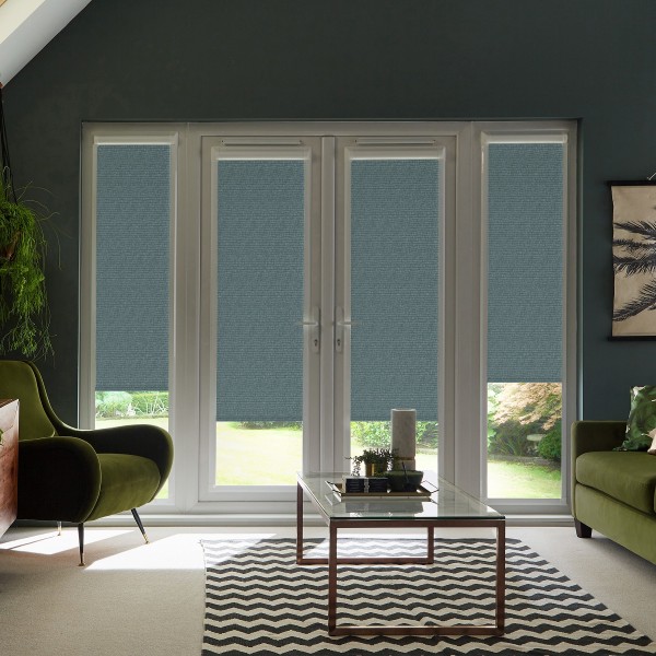 French glass doors with blue roller blinds lowered about two-thirds, framing a sunlit garden beyond; modern living room features green velvet chairs, glass coffee table and chevron-patterned rug.