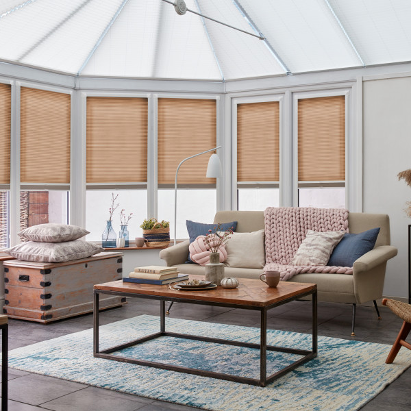 Beige pleated cellular blinds lowered to mid-height across tall conservatory windows, filtering soft daylight; cozy sunroom living area with a neutral sofa, wooden coffee table, pastel throws and blue rug.
