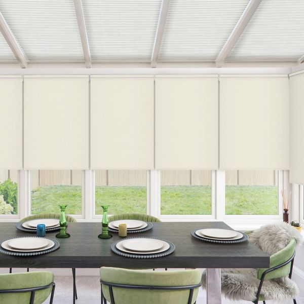 Dining table set with four place settings and decorative glass candlesticks sits beneath lowered cream roller blinds, surrounded by green upholstered chairs and windows revealing a grassy backyard.