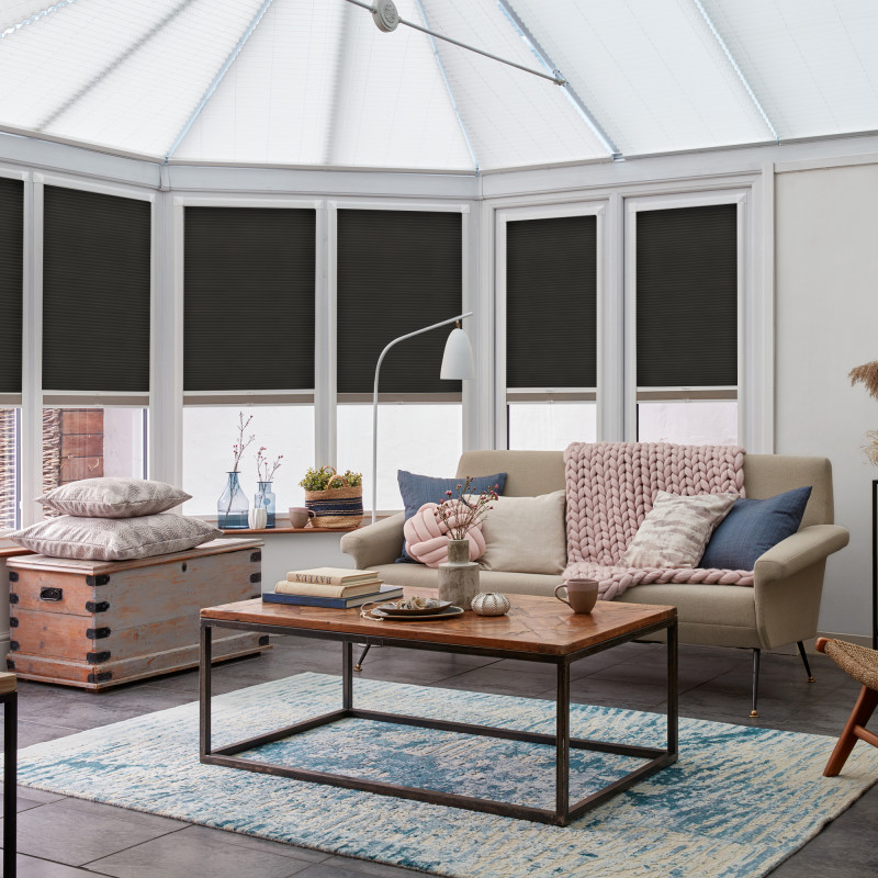Black pleated cellular blinds lowered across conservatory windows, filtering and blocking light; in a bright living area with a beige sofa, chunky pink throw, wooden coffee table and blue patterned rug.