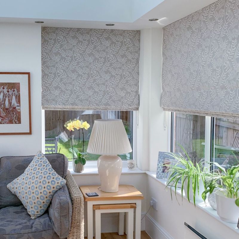 Fabric leaf-patterned Roman blinds, lowered halfway and filtering soft daylight; a sunlit bay living room with a sofa, side table and lamp, orchid and potted plants on the windowsill.