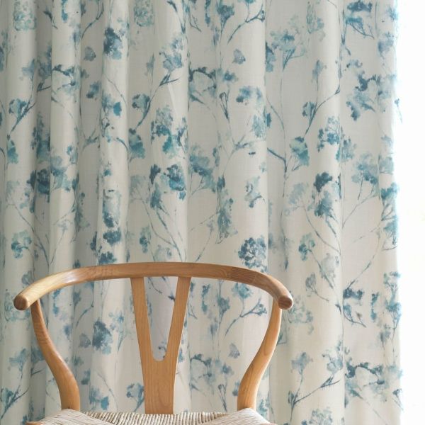 Curtains: full-length fabric panels with watercolor teal-blue floral branches, hanging in soft vertical pleats and drawn closed, gently filtering daylight; context: light-filled room with a wooden wishbone chair foreground.