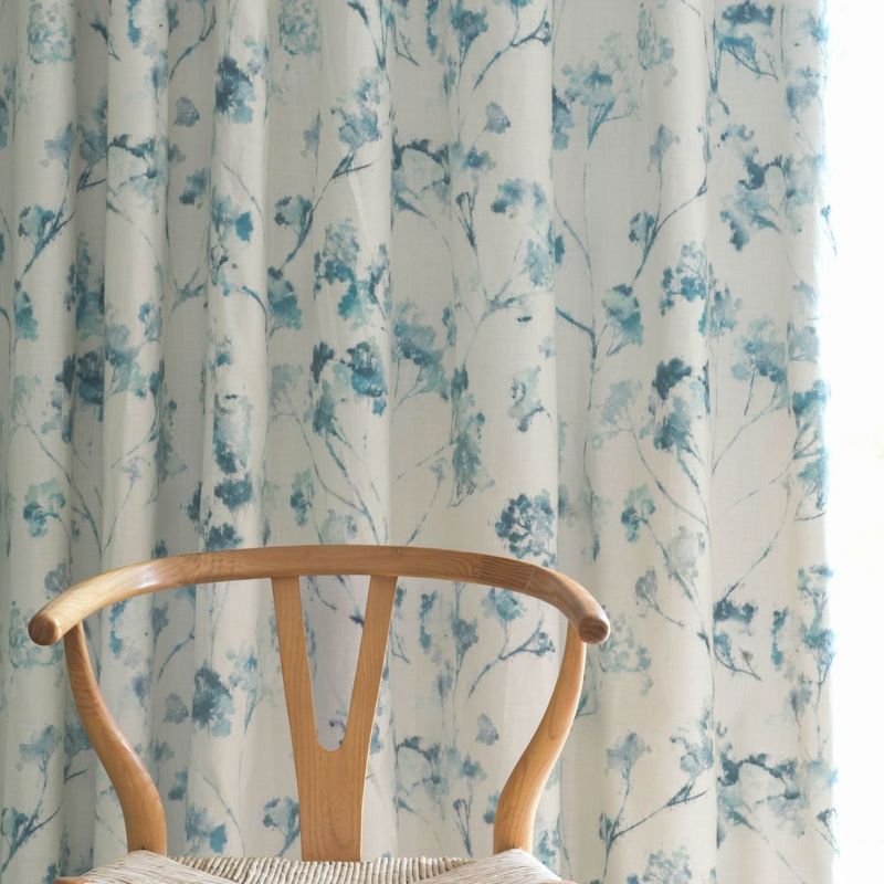 Curtains: full-length fabric panels with watercolor teal-blue floral branches, hanging in soft vertical pleats and drawn closed, gently filtering daylight; context: light-filled room with a wooden wishbone chair foreground.