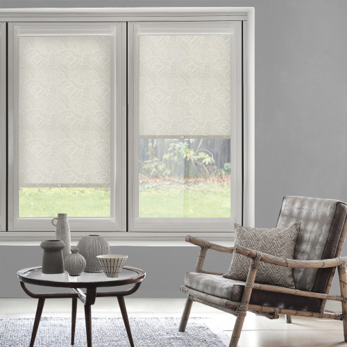 Patterned fabric roller blinds, both mounted inside a double casement frameâ€”left fully lowered, right halfwayâ€”filter daylight; living room with gray walls, rattan armchair, small coffee table, grassy backyard view.