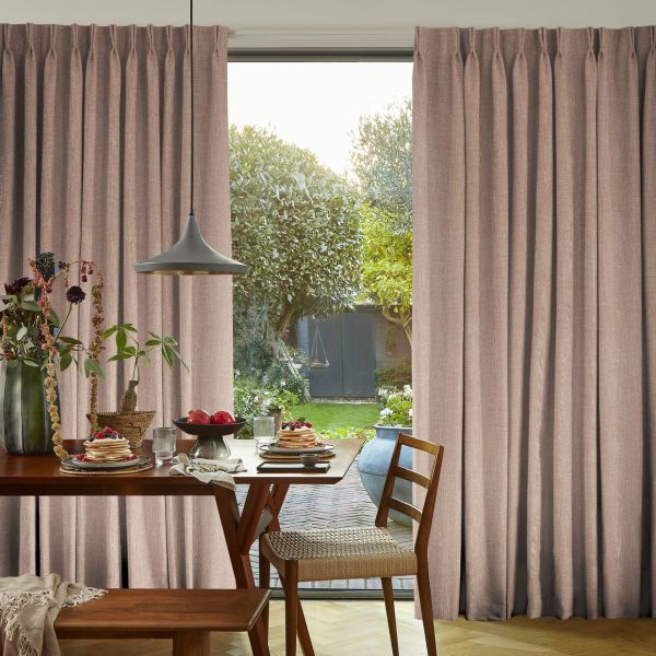 Floor-to-ceiling pleated beige-pink curtains, drawn partially aside to reveal a glass door; curtains filtering daylight. Dining table with pancakes in a warmly lit kitchen opening onto a green garden.