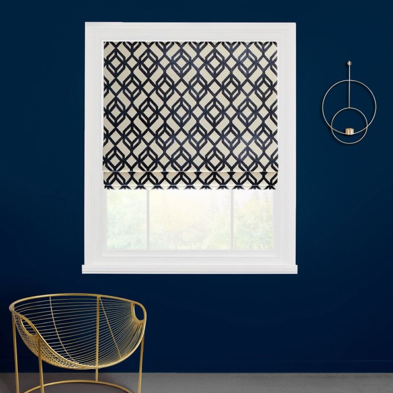 Roman shade with navy geometric lattice, lowered halfway and folding, partially filtering daylight; white-framed window on a deep navy wall, with a gold wire chair and circular gold wall decor.