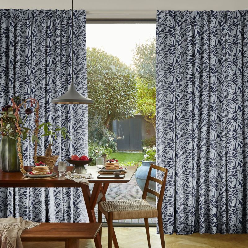 Navy-and-white leaf-patterned pleated curtains, parted to frame a door opening. They reveal sunlit backyard with trees and shed; foreground shows a wooden dining table set for breakfast under pendant lamp.
