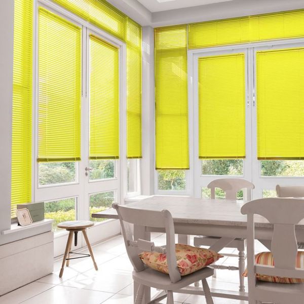Yellow pleated blinds cover tall French doors and windows, lowered and filtering daylight into a sunlit dining nook with a wooden table, chairs, and a garden view.