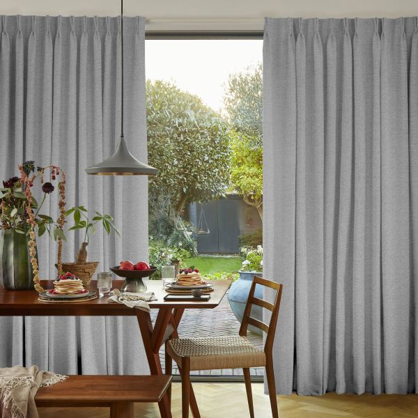 Grey pleated floor-to-ceiling curtains, parted at center in soft folds, filtering daylight; dining table with chairs and breakfast plates inside, backyard garden with trees and a swing visible outside.