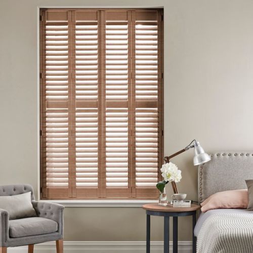 Wooden plantation shutters â€” four full-height panels with horizontal louvers â€” cover a recessed window, closed and angling slats to filter daylight in a neutral bedroom with armchair, bed, side table.