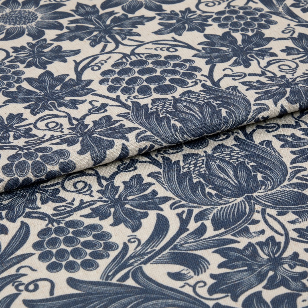 Blue floral-and-grape patterned upholstery fabric, folded to show thickness and weave, printed in indigo on a natural beige ground; close-up, studio-lit textile surface.