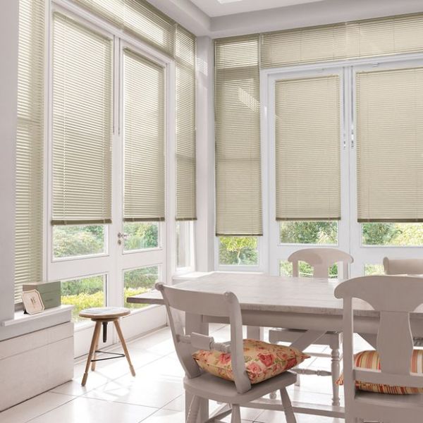 Beige horizontal Venetian blinds covering tall bay windows, lowered and filtering soft daylight; in a bright sunroom dining area with white table, chairs, floral cushions and garden view.