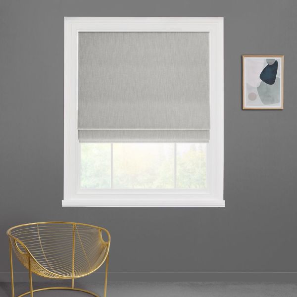 Grey Roman shade lowered with folded hem, partially filtering daylight; white-trimmed window set in a dark grey wall with a gold wire chair and small abstract framed art, blurred greenery.