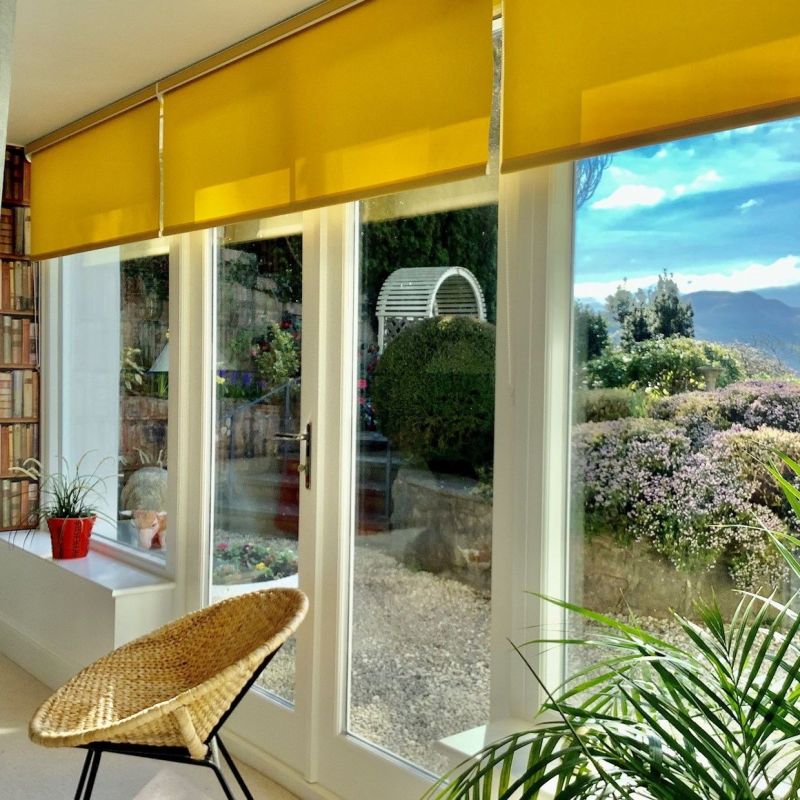Yellow roller blinds across the top of glass patio doors, partially lowered and filtering sunlight; wicker chair and potted plants inside, gravelled stone garden and distant hills visible outside.
