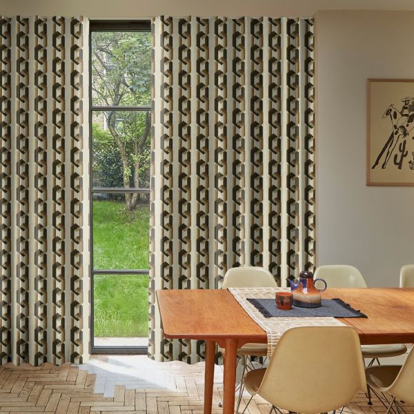 Geometric-print, full-length curtains with a linked-chain motif, hung floor-to-ceiling and parted to frame a narrow central window, filtering daylight into a mid-century dining room with wooden table and garden view.