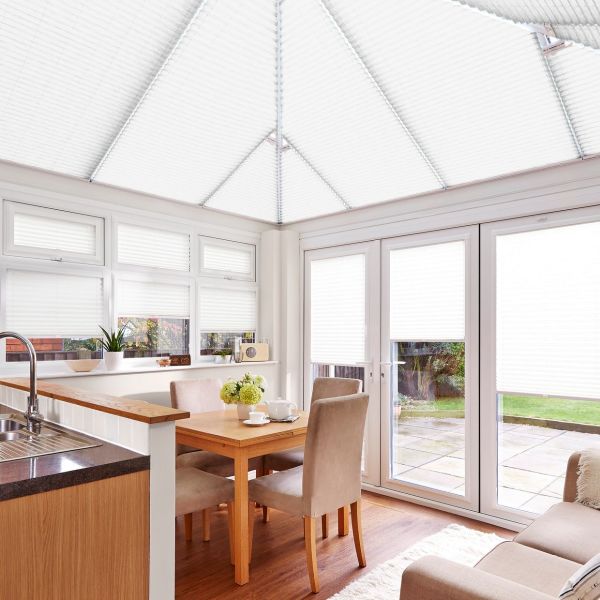 White pleated cellular blinds covering conservatory roof, windows and patio doors, fully lowered and softly filtering daylight; small dining area with wooden table and upholstered chairs, bright, garden view.