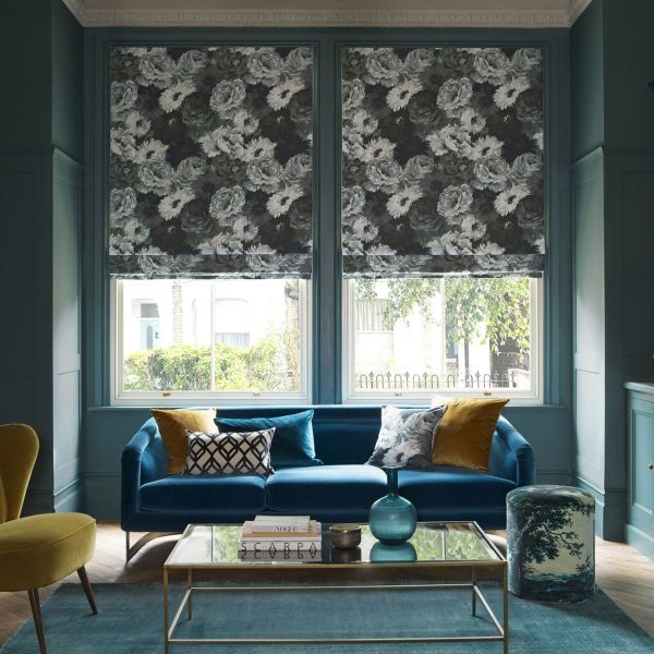 Floral Roman blinds over two tall windows, lowered and patterned with large monochrome roses, filtering daylight into a teal living room with a blue velvet sofa and glass coffee table.
SCARPA
