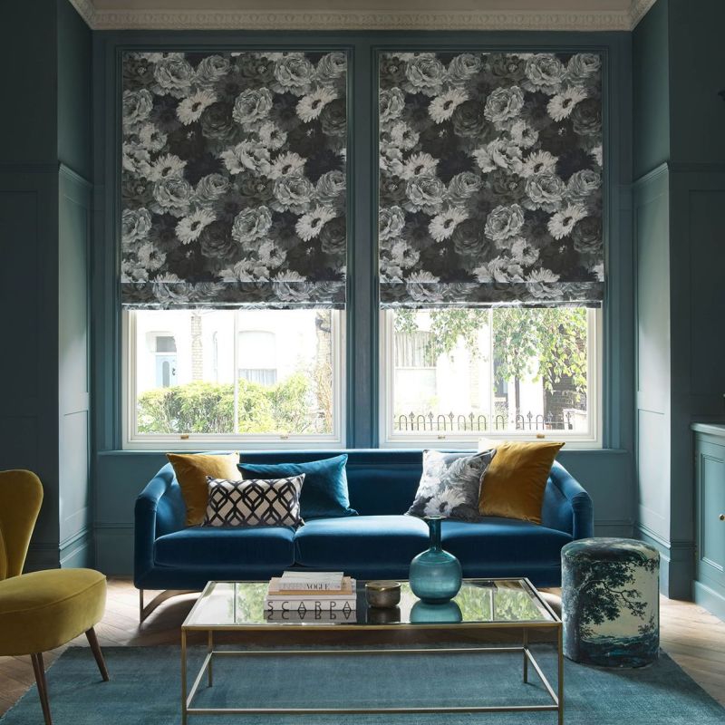 Floral Roman blinds over two tall windows, lowered and patterned with large monochrome roses, filtering daylight into a teal living room with a blue velvet sofa and glass coffee table.

SCARPA