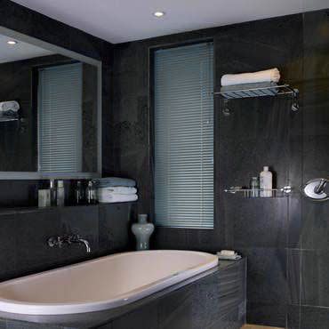 Narrow vertical venetian blinds covering a tall bathroom window, closed and filtering soft daylight; dark slate-tiled walls, freestanding tub, mirrored shelf, towels and toiletries nearby.