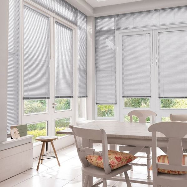White horizontal blinds covering tall bay windows, lowered with thin slats partially closed, filtering soft daylight; pale dining table and chairs in bright breakfast nook facing a green garden.